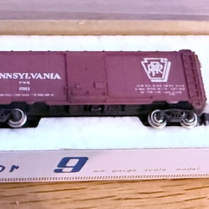 May include: A brown boxcar model train car with the words "Pennsylvania RR" and a Pennsylvania state logo printed on the side. The boxcar is on a white background with the words "Con-Cor 9" printed on the bottom.