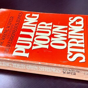 May include: A hardcover book titled "Pulling Your Own Strings" by Dr. Wayne W. Dyer. The book has a bright orange cover with white lettering. The book's title is prominently displayed, along with the author's name and a subtitle.