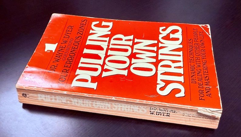 May include: A hardcover book titled "Pulling Your Own Strings" by Dr. Wayne W. Dyer. The book has a bright orange cover with white lettering. The book's title is prominently displayed, along with the author's name and a subtitle.