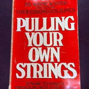 May include: A vintage paperback book titled "Pulling Your Own Strings" by Dr. Wayne W. Dyer. The cover is red with white lettering. The book's title is prominently displayed, along with the subtitle and author's name.