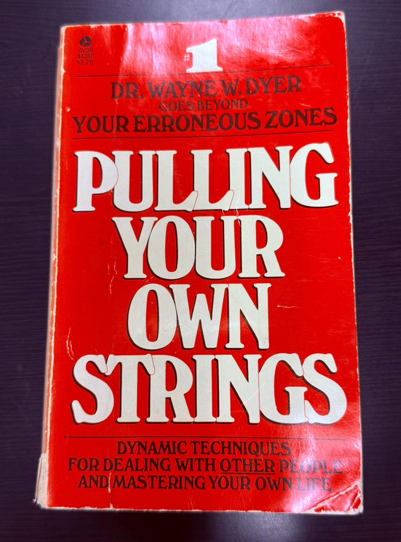 May include: A vintage paperback book titled "Pulling Your Own Strings" by Dr. Wayne W. Dyer. The cover is red with white lettering. The book's title is prominently displayed, along with the subtitle and author's name.