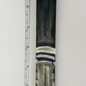 includes : A handcrafted V for Vendetta-themed letter opener. The top features a sculpted V for Vendetta character head, black hat, and coat. The handle is painted black with white stripes, transitioning to a gray and black blade. The letter opener measures approximately 9 inches tall, as shown next to a measuring tape.