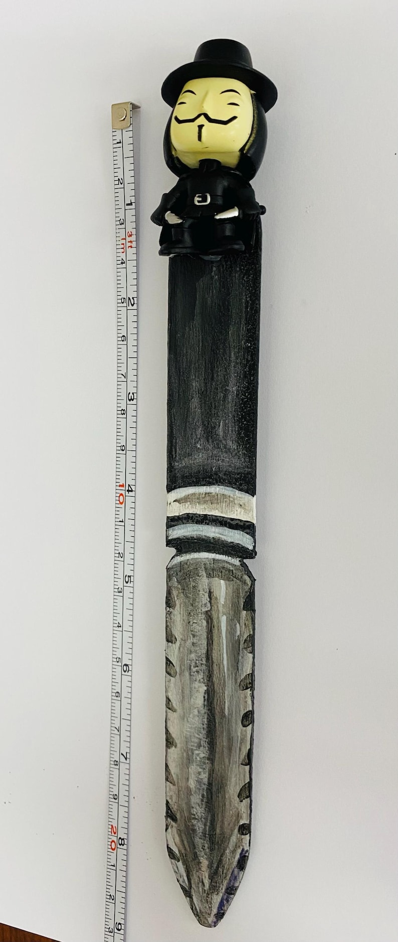 includes : A handcrafted V for Vendetta-themed letter opener. The top features a sculpted V for Vendetta character head, black hat, and coat. The handle is painted black with white stripes, transitioning to a gray and black blade. The letter opener measures approximately 9 inches tall, as shown next to a measuring tape.
