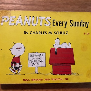 May include: A yellow book cover with the title "PEANUTS Every Sunday" by Charles M. Schulz. The cover features a cartoon illustration of Charlie Brown walking and Snoopy sleeping on his doghouse.
