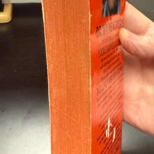 May include: A vintage paperback book with an orange cover and spine. The book's title is visible, along with the author's name and a portrait. The book is held by a hand.