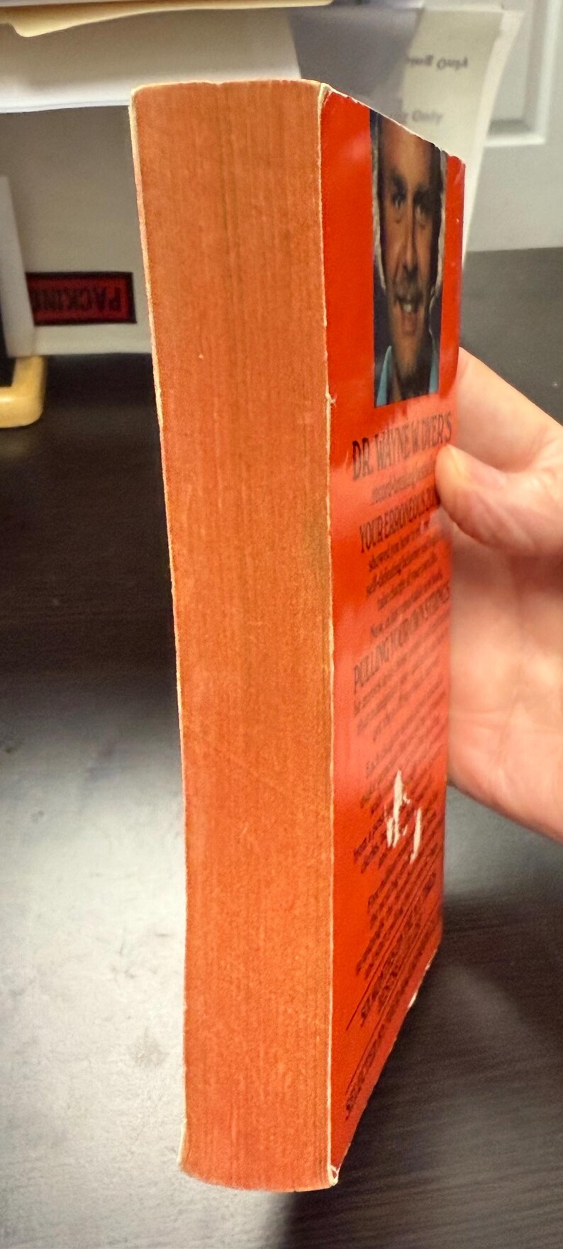 May include: A vintage paperback book with an orange cover and spine. The book's title is visible, along with the author's name and a portrait. The book is held by a hand.