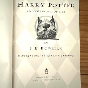 May include: Open book with the title "Harry Potter and the Goblet of Fire" by J.K. Rowling. The book's cover features an illustration of a goblet with flames, set against a diamond pattern background. The book is published by Arthur A. Levine Books.