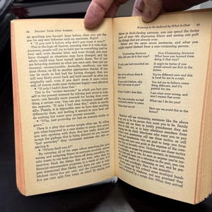May include: Open book with text on pages 62 and 63. The text discusses victimizing and non-victimizing sentences, and how to avoid fault-finding patterns. The book is open in a person's hand.