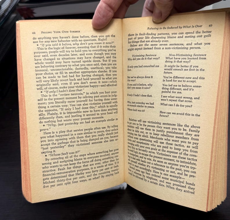May include: Open book with text on pages 62 and 63. The text discusses victimizing and non-victimizing sentences, and how to avoid fault-finding patterns. The book is open in a person's hand.