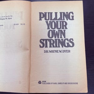 May include: Open book with the title "PULLING YOUR OWN STRINGS" by Dr. Wayne W. Dyer. The book is published by Avon. The cover is a light tan color with black text.