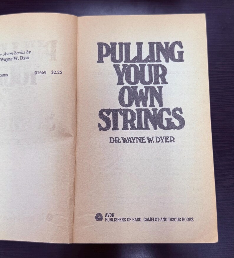 May include: Open book with the title "PULLING YOUR OWN STRINGS" by Dr. Wayne W. Dyer. The book is published by Avon. The cover is a light tan color with black text.