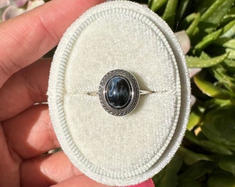Op de afbeelding: Een zilveren ring met een ovale zwarte en witte edelsteen gezet in een zilveren bezel. De ring ligt op een wit fluwelen ringendoosje.