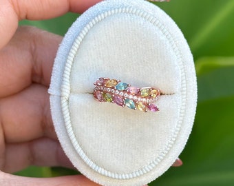 Può includere: Un anello in oro rosa con un design a foglia, adornato da una fila di pietre preziose colorate e piccoli diamanti. L'anello è presentato su un portagioie in velluto bianco, con foglie verdi sullo sfondo.