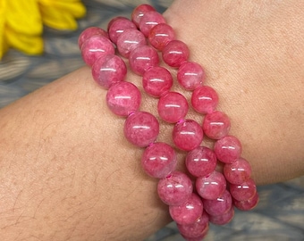 May include: A pink gemstone bracelet with three strands of round beads. The bracelet is worn on a person's wrist.