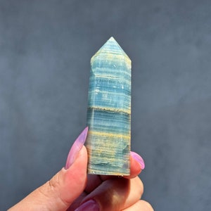 Peut inclure: Une pointe de cristal bleu et jaune, également appelée baguette de cristal, est tenue dans une main avec un vernis à ongles rose.