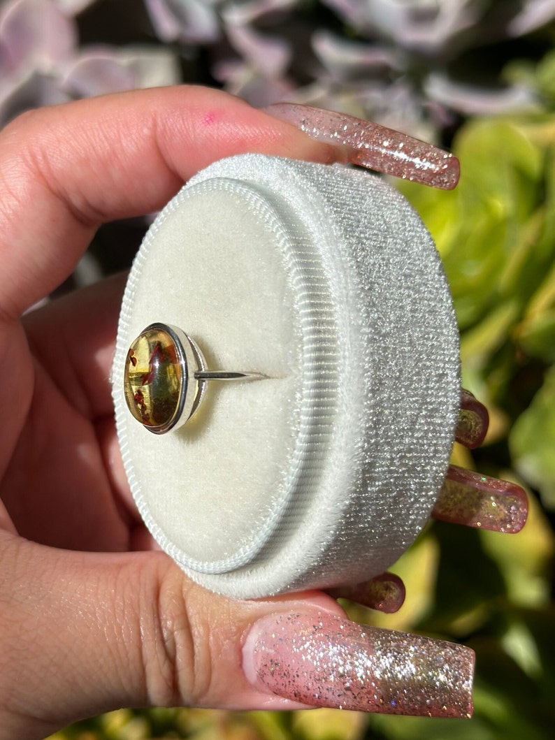 Op de afbeelding: Een zilveren ring met een ronde amber steen gezet in een bezel setting. De steen is diep oranje van kleur met vlekken van bruin en geel. De ring wordt getoond op een wit fluwelen ringdoosje.