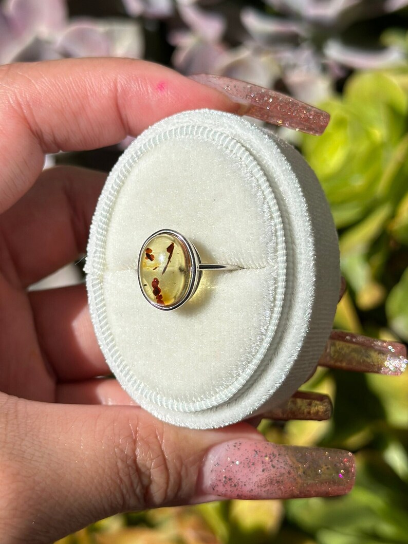 Op de afbeelding: Een zilveren ring met een gele ovale barnsteen in een bezel setting. De barnsteen heeft kleine bruine insluitsels.