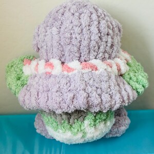May include: A handmade, plush toy in the shape of a bear, viewed from the back. The bear is primarily lavender, with green and white accents on the arms and bottom. A white and pink striped collar is around the neck. The toy is made of soft, textured yarn.