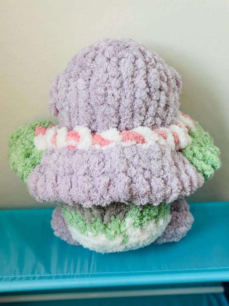 May include: A handmade, plush toy in the shape of a bear, viewed from the back. The bear is primarily lavender, with green and white accents on the arms and bottom. A white and pink striped collar is around the neck. The toy is made of soft, textured yarn.