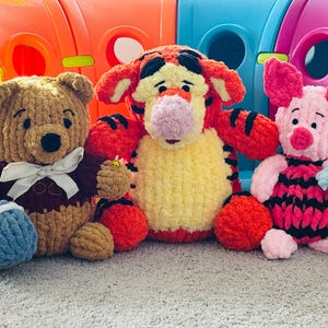 May include: Three handmade crochet plush toys featuring a brown bear holding a blue honey pot, a bright orange and yellow tiger with black stripes, and a pink piglet with black stripes holding a small blue and white flower with green leaves.