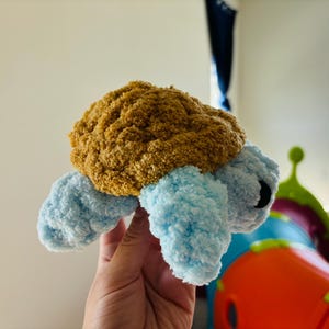 May include: A handmade plush turtle with a brown shell and light blue flipper-like limbs. The turtle has black eyes and is made of soft, textured yarn. This cuddly toy is perfect for children.