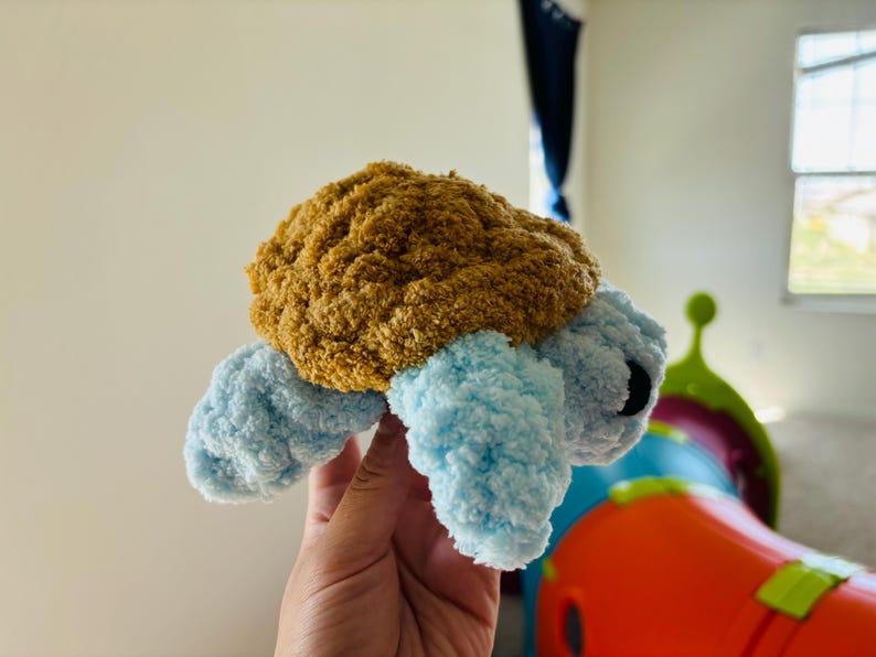May include: A handmade plush turtle with a brown shell and light blue flipper-like limbs. The turtle has black eyes and is made of soft, textured yarn. This cuddly toy is perfect for children.