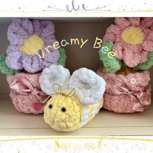 May include: A handmade, plush bee with white wings and a yellow and white body sits in front of two flower-shaped plushies. The flowers are pink and purple with green leaves and yellow centers. The text "dreamy Bee" is in the center.