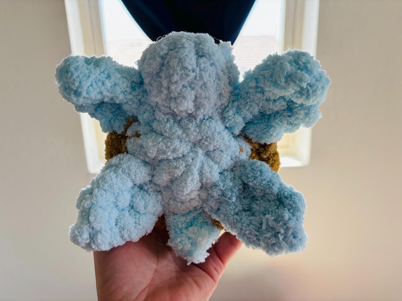 May include: A handmade, plush, blue and brown crocheted flower. The flower has large, fluffy petals in shades of light blue, with a brown center. The soft texture and vibrant colors make it a charming decorative item.