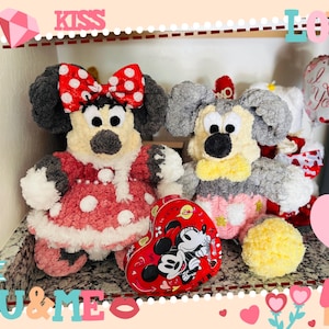 May include: Two plush toys, one a grey Mickey Mouse with a yellow bow tie and the other a red and white Minnie Mouse with a red bow, are sitting on a shelf with a heart-shaped box of chocolates. The box has a picture of Mickey and Minnie Mouse on it. The shelf is decorated with hearts and the words "Kiss", "Love", and "You & Me".