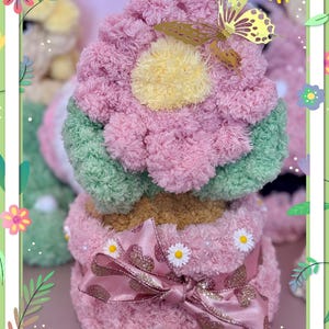 Może przedstawiać: Różowy, zielony i żółty prezent w kształcie kwiatu z dekoracyjną kokardą i złotym motylem. Prezent jest ozdobiony małymi białymi stokrotkami i napisem "Happy Mother's Day".