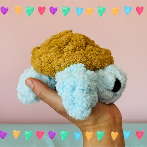 May include: A handmade plush turtle toy with a light blue body and a textured, golden-brown shell. The turtle has black eyes and is held in a hand. The background is a soft pink with a border of colorful hearts.