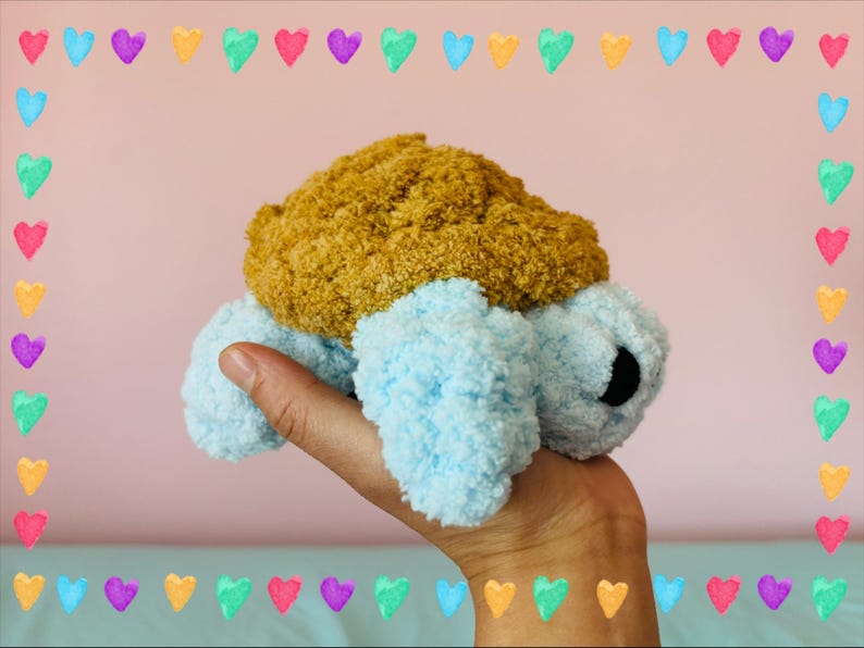 May include: A handmade plush turtle toy with a light blue body and a textured, golden-brown shell. The turtle has black eyes and is held in a hand. The background is a soft pink with a border of colorful hearts.