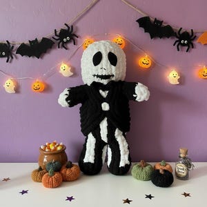 May include: A plush crochet doll featuring a white round head with black stitched eyes and a wide stitched mouth, wearing a black and white pinstriped suit with a black bowtie. The doll is surrounded by small crocheted pumpkins and a glass bottle.