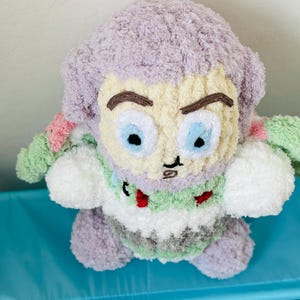 May include: A handmade plush toy of a cartoon character with a light purple head and hair, a cream-colored face, and blue eyes. The toy has a green and white body with red accents. The toy is sitting on a blue surface.