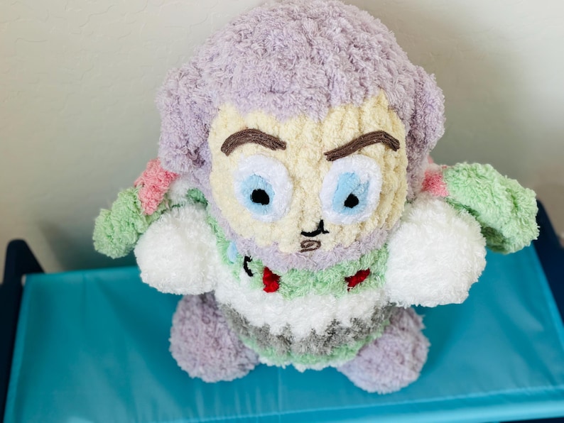 May include: A handmade plush toy of a cartoon character with a light purple head and hair, a cream-colored face, and blue eyes. The toy has a green and white body with red accents. The toy is sitting on a blue surface.