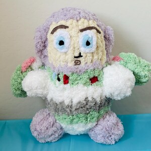 May include: A handmade plush toy resembling a cartoon character. The toy features a light purple head and limbs, a cream-colored face with blue eyes, and a body with green, white, and gray stripes. Small red accents are visible on the chest.