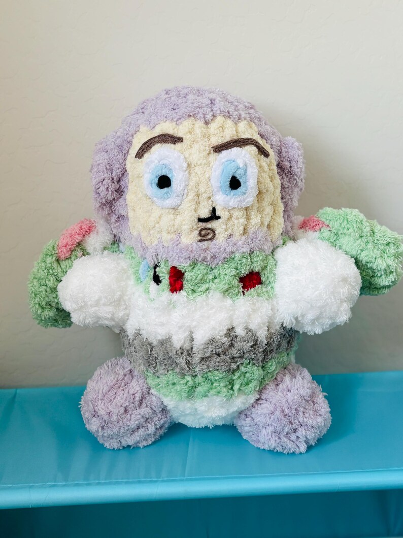 May include: A handmade plush toy resembling a cartoon character. The toy features a light purple head and limbs, a cream-colored face with blue eyes, and a body with green, white, and gray stripes. Small red accents are visible on the chest.
