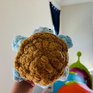 May include: A handmade plush turtle toy with a brown shell and light blue flippers and head. The turtle is made of soft, textured yarn, and is held in a person's hand. The toy is designed for children.