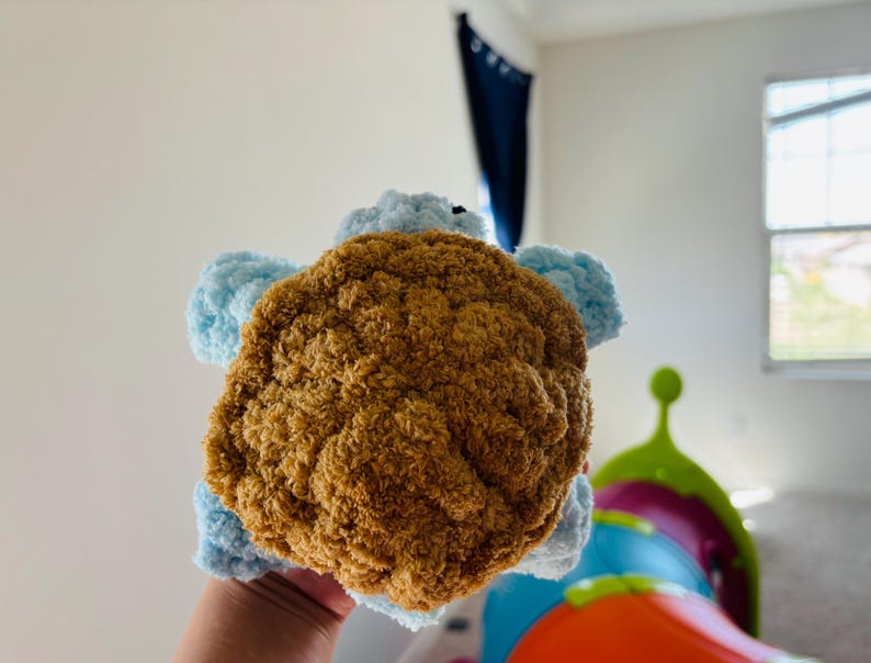 May include: A handmade plush turtle toy with a brown shell and light blue flippers and head. The turtle is made of soft, textured yarn, and is held in a person's hand. The toy is designed for children.