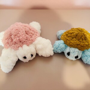 May include: Two plush turtle toys. One has a pink shell, white flippers, and black eyes. The other has a mustard-colored shell, blue flippers, and black eyes. Both are made of soft, fluffy material.