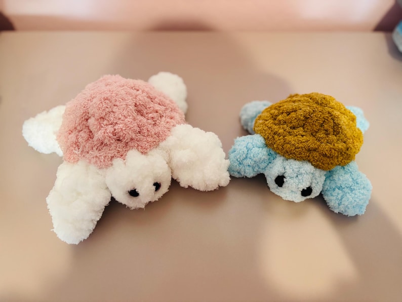 May include: Two plush turtle toys. One has a pink shell, white flippers, and black eyes. The other has a mustard-colored shell, blue flippers, and black eyes. Both are made of soft, fluffy material.