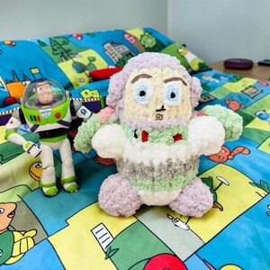 May include: A plush, handmade toy in the shape of a character from the movie Toy Story, with a Buzz Lightyear action figure to the left. The plush toy has a light purple head, blue eyes, and a white and green body. The background is a blue patterned bedspread.