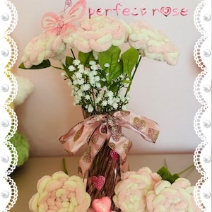 May include: A bouquet of pink and white finger-knitted roses in a vase with a gold ribbon and heart-shaped embellishments. The text "Finger knit the perfect rose" is displayed above the bouquet.