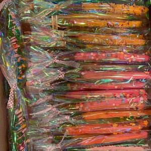 Puede incluir: Una variedad de palitos de pretzel cubiertos de chocolate y espolvoreados con coloridos sprinkles, empaquetados individualmente en bolsas de celofán transparentes con cintas de lunares rojos y blancos.