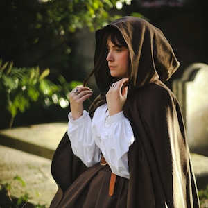 Peut inclure: Une femme portant une cape à capuche marron avec une chemise blanche et une jupe marron. Elle se tient dans un cimetière avec de la végétation verte en arrière-plan.