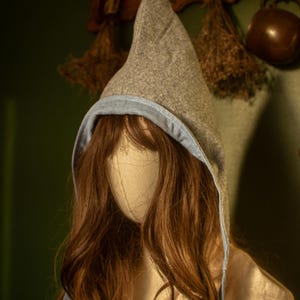 Peut inclure: Une cape à capuche grise avec une pointe, doublée de tissu bleu clair. Le capuchon est présenté sur une tête de mannequin avec de longs cheveux bruns ondulés. L'arrière-plan comprend des herbes séchées et un mur vert foncé.