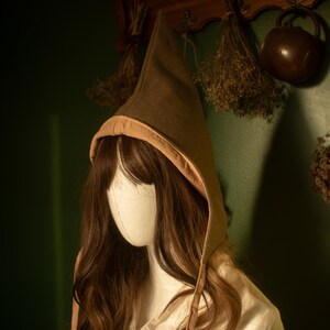 Peut inclure: Un chapeau à capuche marron avec un sommet pointu et une bordure beige. Le chapeau est présenté sur une tête de mannequin avec de longs cheveux bruns, portant un vêtement crème. L'arrière-plan présente un mur vert foncé et des accents en bois.