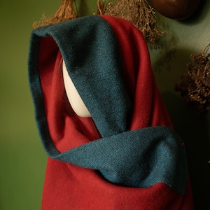 Może przedstawiać: Czerwony płaszcz z turkusowym kapturem. Kaptur ma wzór w romby. Płaszcz jest narzucony na białą głowę manekina. Tło to zielona ściana z suszonymi ziołami i brązowym garnkiem.