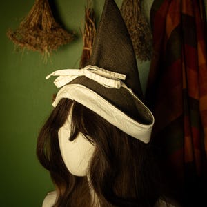 May include: A dark brown witch hat with a white brim and bow, displayed on a mannequin head with long brown hair. The hat has a pointed crown and a wide brim. The background includes dried herbs and a plaid textile.