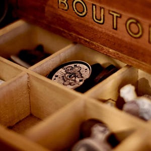 May include: A wooden box with the word "BUTTONS" on the lid. The box is open and contains various buttons, including a round button with a black and white eye design.